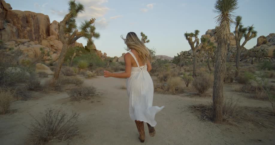 A woman with long blonde hair and a white dress runs on a dirt path in a desert landscape surrounded by joshua trees and large rock formations