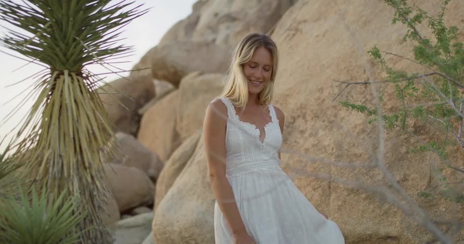 A woman in a white dress stands near a joshua tree and large rocks touching her hair