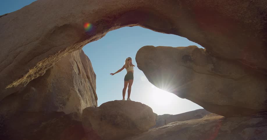 A young woman is standing under a large rock archway in a desert landscape she is silhouetted against the bright sky as she stretches her arms above her head and then lowers them