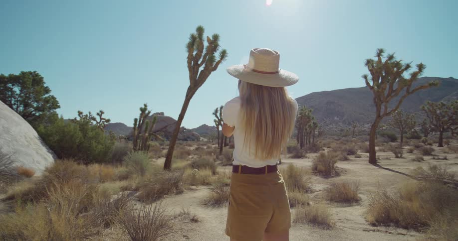 Woman wearing a straw hat photographs desert landscape with joshua trees