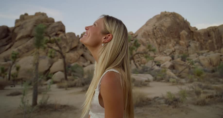 A blonde woman in a white dress and pearl earrings looks up and smiles in a desert landscape with rocky formations and joshua trees