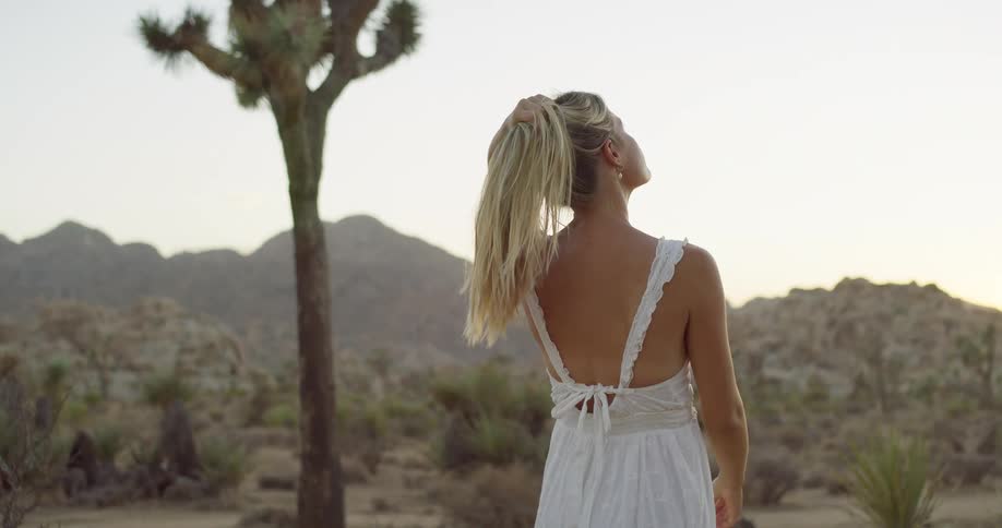 A woman wearing a long white dress and brown cowboy boots walks on a sandy desert path with joshua trees and hills in the background facing away from the camera before turning and smiling at the camera