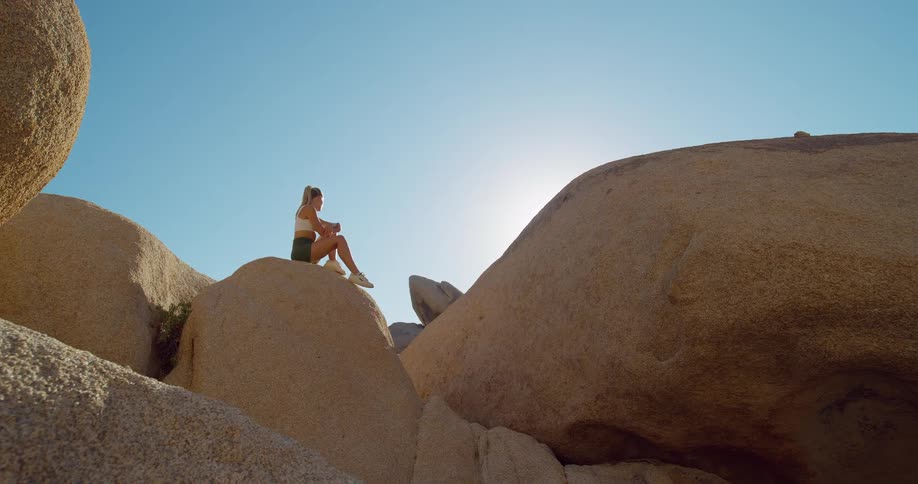 A young woman wearing short activewear sits on top of a rock in a rocky formation at midday