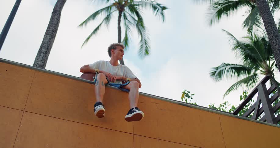 The video shows a lowangle shot of a young man with a skateboard sitting on a wooden ledge outdoors he is looking off into the distance with palm trees visible behind him against a bright sky