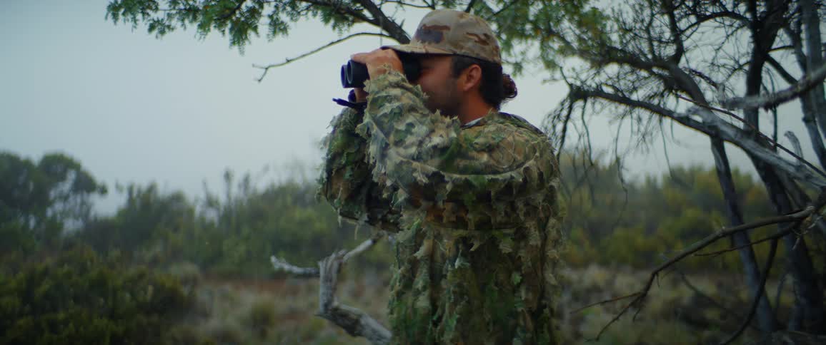 A man wearing camouflage looks through a pair of binoculars