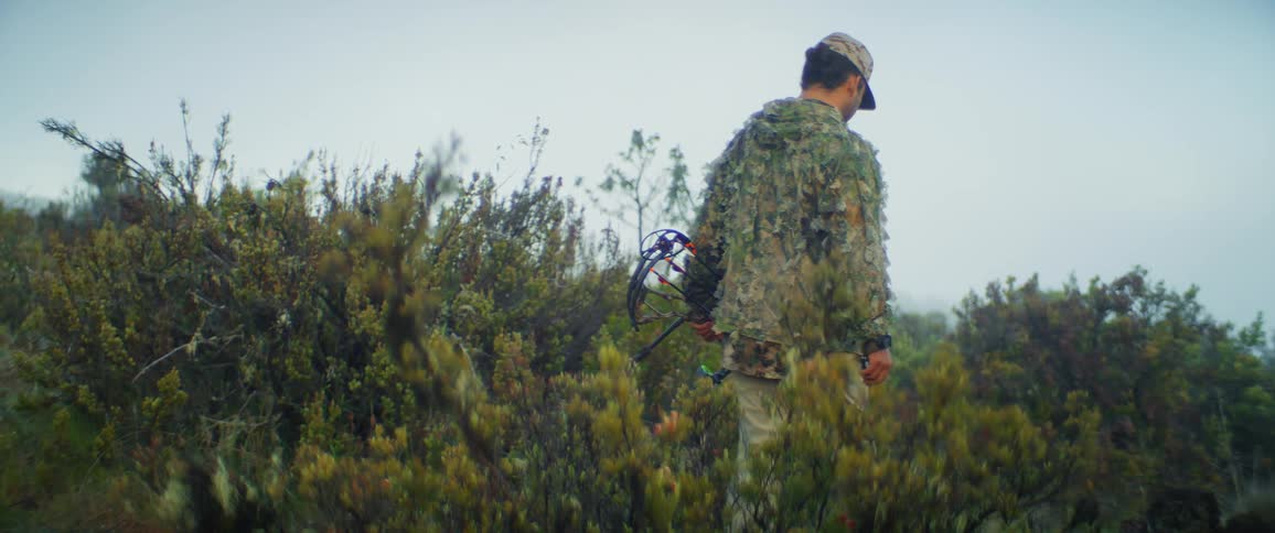 Man in camouflage clothing walks among bushes carrying a compound bow