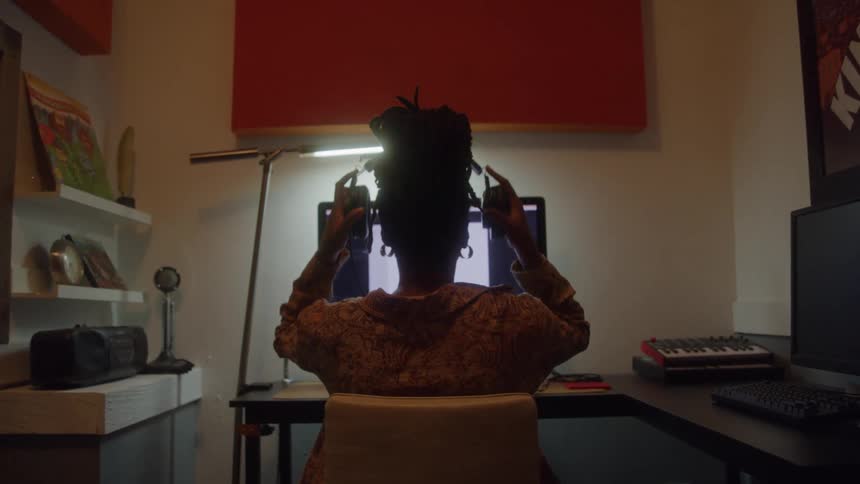 A person with long hair tied up sits at a desk in a recording studio they are facing a computer screen and putting on a pair of headphones
