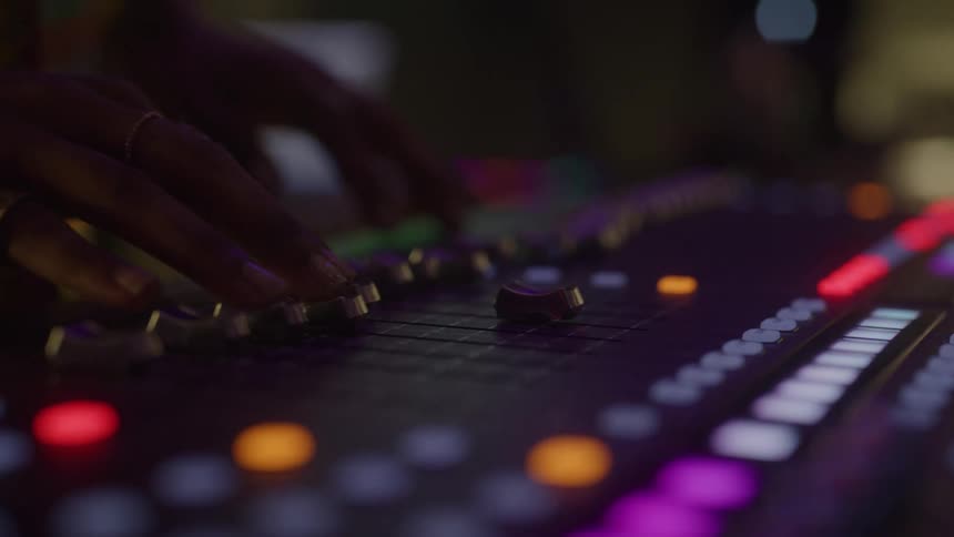 The video shows a persons hands operating a sound mixing board the hands are moving the faders and knobs which are lit up with bright colorful lights