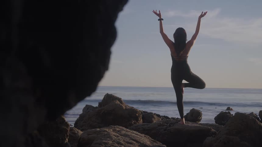 A woman balances on one leg while doing yoga overlooking the ocean