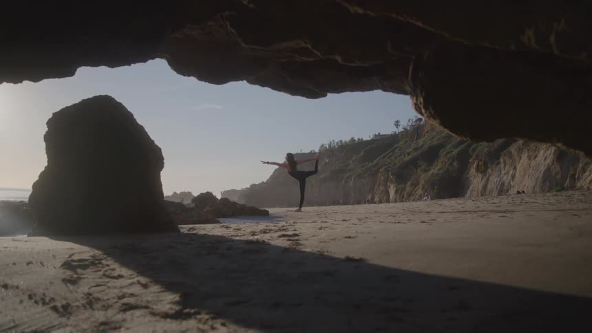 A young woman doing yoga beside a cave on the beach at sunset
