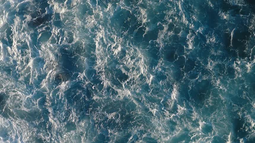 A topdown view captures churning ocean waves white foam lacing across deep blue water in intricate patterns