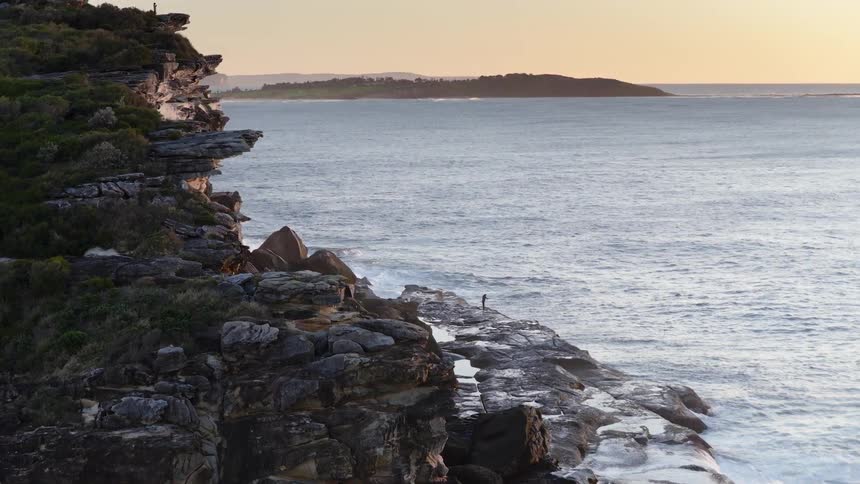 Rocky cliff by the sea at sunset with a person standing near the waves
