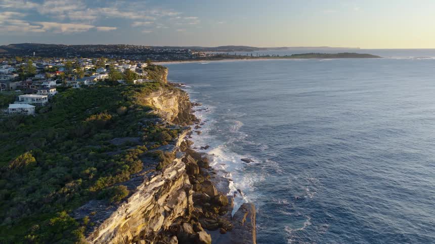 A wide aerial view of a dramatic coastline with waves crashing against a rugged cliff face a town perched on the edge and the ocean stretching out to the horizon under a soft sky