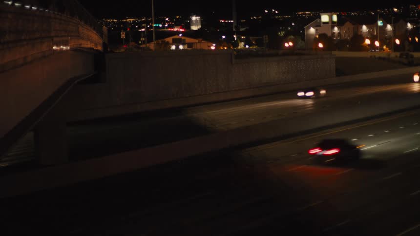A view from above an overpass shows cars driving on a multilane highway at night with the bright headlights and taillights creating light streaks and a city visible in the distance