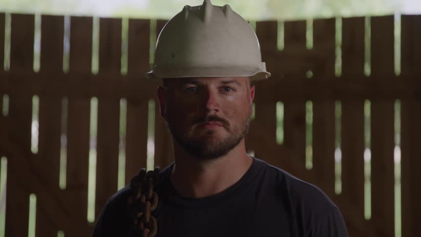 A man with a light beard is standing inside a structure with wooden beams he is wearing a dark tshirt and a white hard hat a heavy metal chain is draped over his shoulder