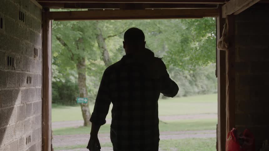 A man seen in silhouette is walking away from the camera and out of a dark barn carrying a tool over his shoulder and a cloth in his hand towards a bright green outdoor field