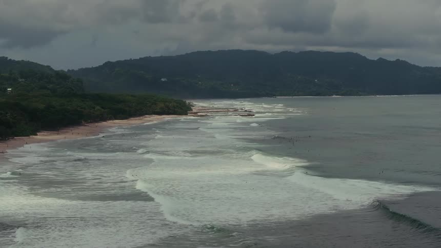An aerial view shows powerful white waves rolling onto a sandy beach with a lush green shoreline leading up to large cloudy mountains in the background