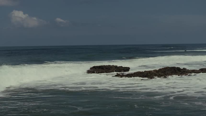 Powerful waves are seen crashing over dark rocks in the middle of a vast ocean under a partly cloudy sky