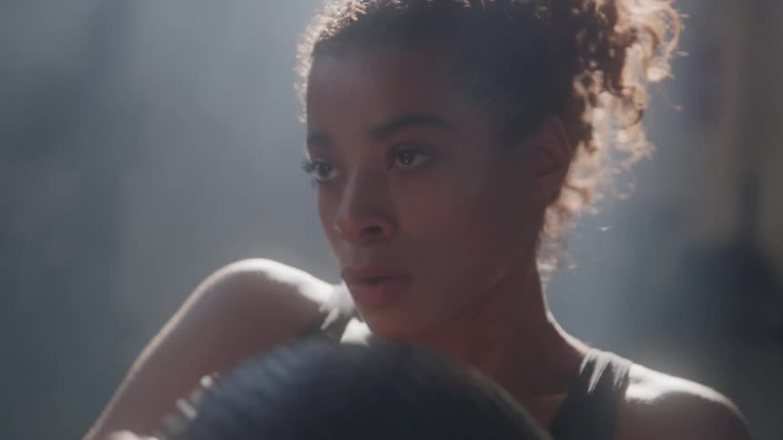 A young woman is shown in a closeup shot looking at something offcamera with a serious expression she is in a gym wearing a black top and her hair is in a messy bun