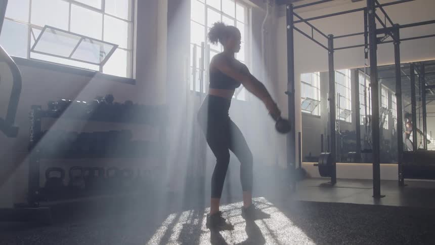 A woman exercises with a kettlebell in a sunlit gym