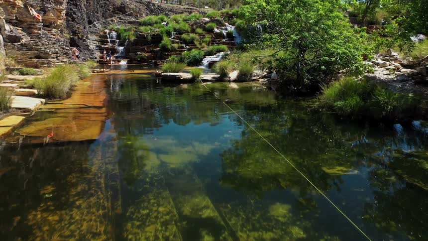 A serene river with clear water is shown with a small waterfall cascading down a rocky green hill in the background the camera pulls back revealing a few people swimming in the water and a lush natural landscape