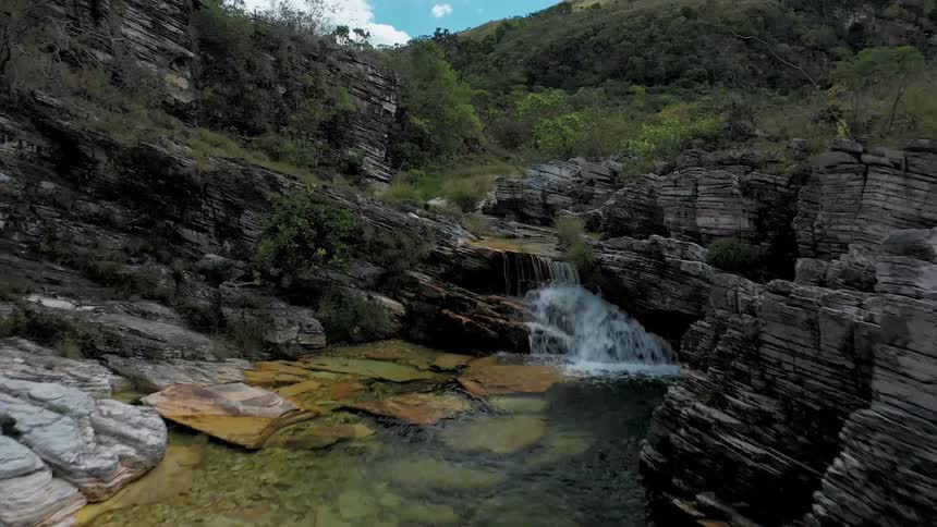 A river with clear water and a small waterfall flows through a canyon of gray and white rocks surrounded by green foliage on the rocky hillsides under a cloudy blue sky