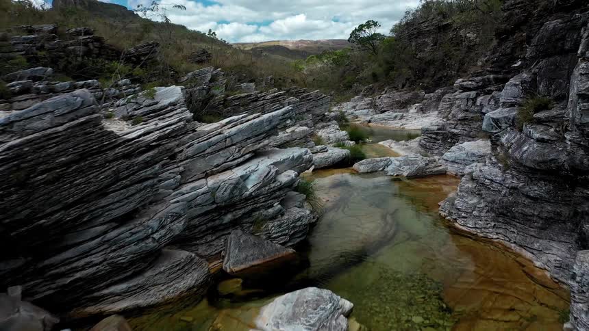 A low aerial view moves forward over a narrow shallow stream flowing through a gorge with light gray layered rock walls and small patches of green grass under a blue sky with white clouds