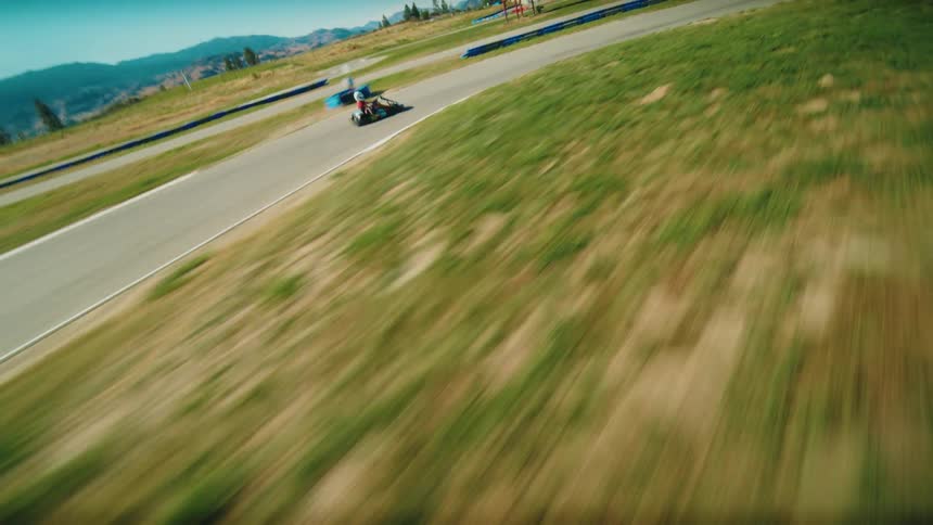A firstperson perspective shows a person in a gokart speeding along a track the camera passes a small white barrier on the left and a gokart in front is visible as they approach a turn