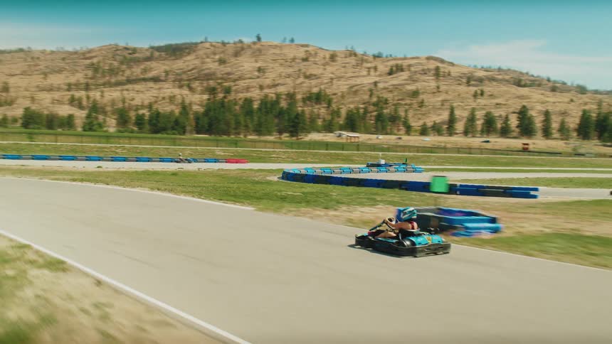 Gokarts are racing on a track in a scenic rural area with mountains in the background the camera pans from left to right following the karts as they speed down the paved course