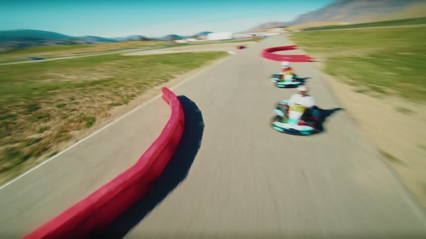 A high angle drone view follows a go kart on a race track as the driver navigates turns while other go karts race nearby with a hilly landscape and clear blue sky in the distance