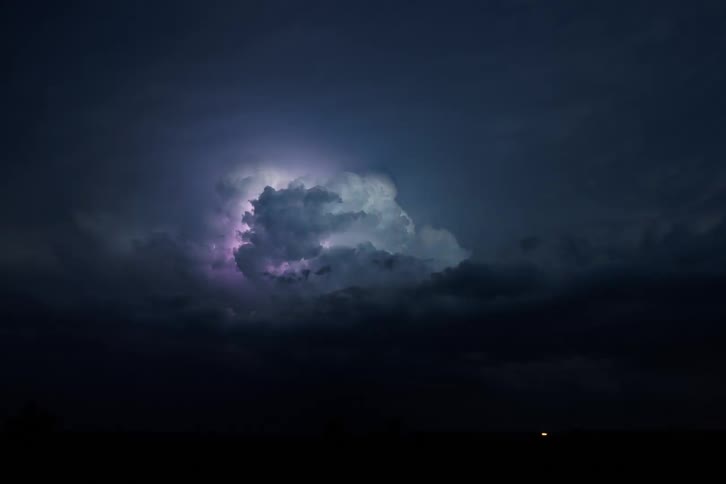 A flash of bright white lightning illuminates a large dark cumulus cloud in the center of the frame surrounded by dark storm clouds and a black horizon