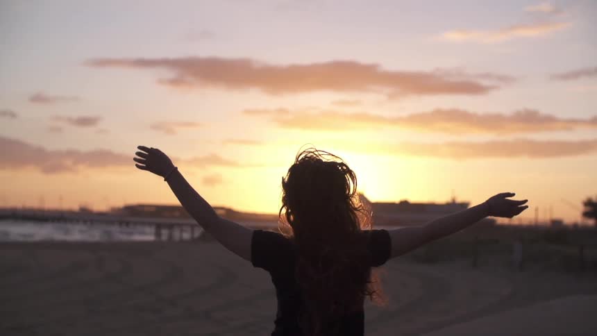 A woman with long hair and outstretched arms running down a street located next to a beach at sunset