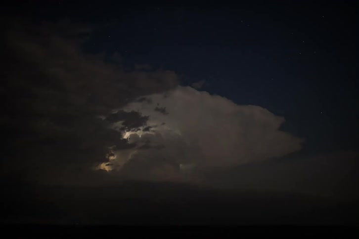 Flashes of lightning illuminate a massive storm cloud from within against a dark starry night sky