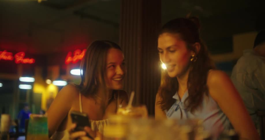 Two young women smile while looking at a phone in a bar lit by neon lights