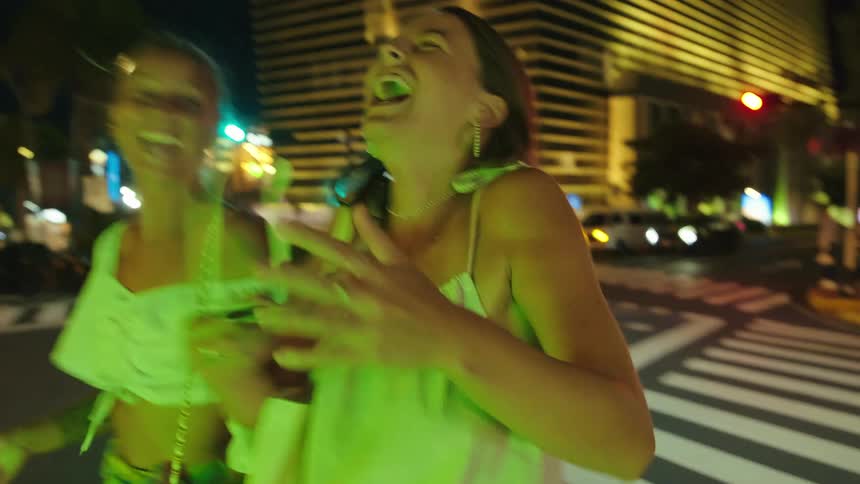 Two white young women walking and laughing while they cross the streets and dance playfully outdoors during night time in a metropolitan area