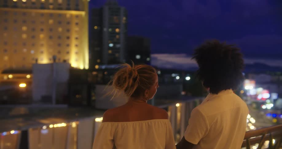 Friends standing on a balcony overlooking the city at night smile and talk