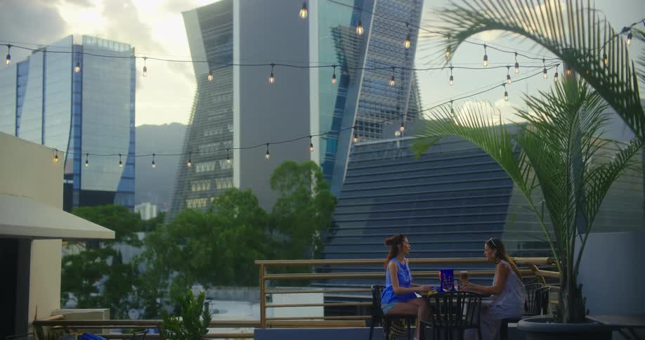 Two women sit at a rooftop cafe with drinks overlooking the city