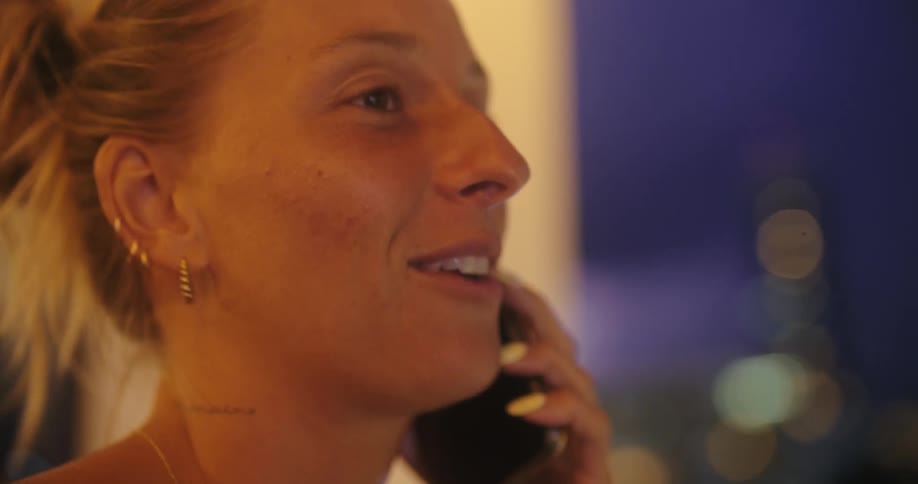 A young blonde woman making a phone call on her hotel room balcony at night laughing during the conversation