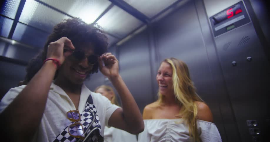A young man takes off his glasses and puts them on a young woman who seems excited as they both laugh while riding an elevator