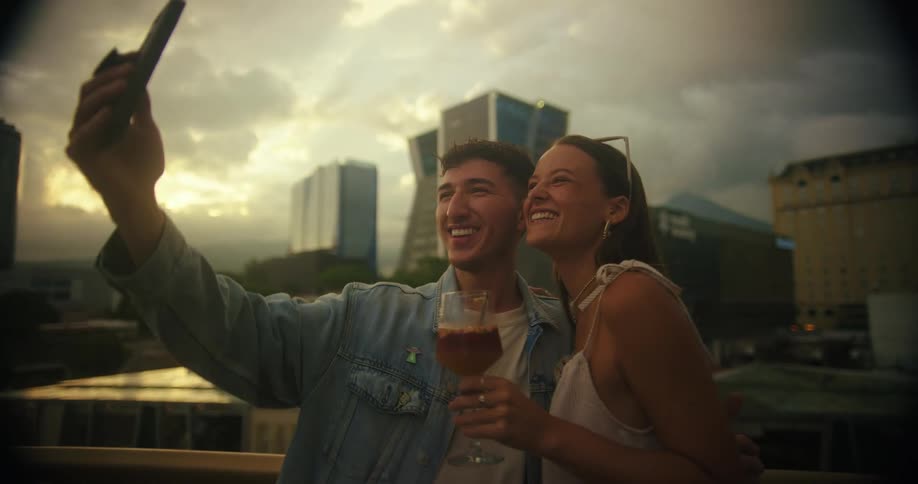 Couple smiles taking a selfie on a rooftop with buildings in the background during sunset