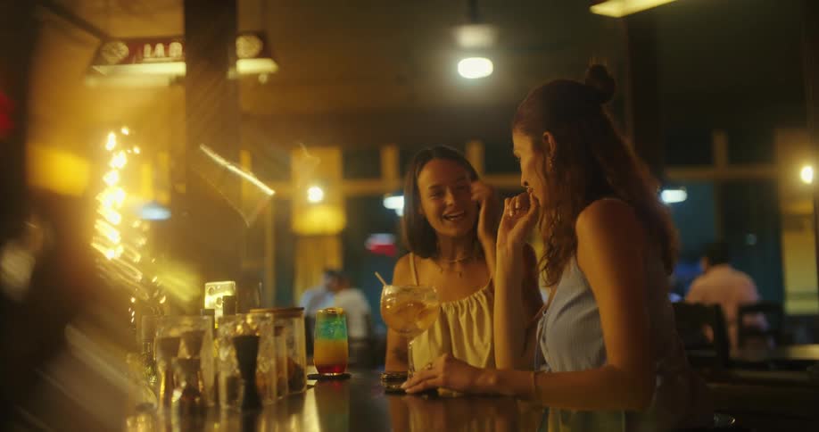 Two women enjoy drinks and laugh together at a cozy bar at night