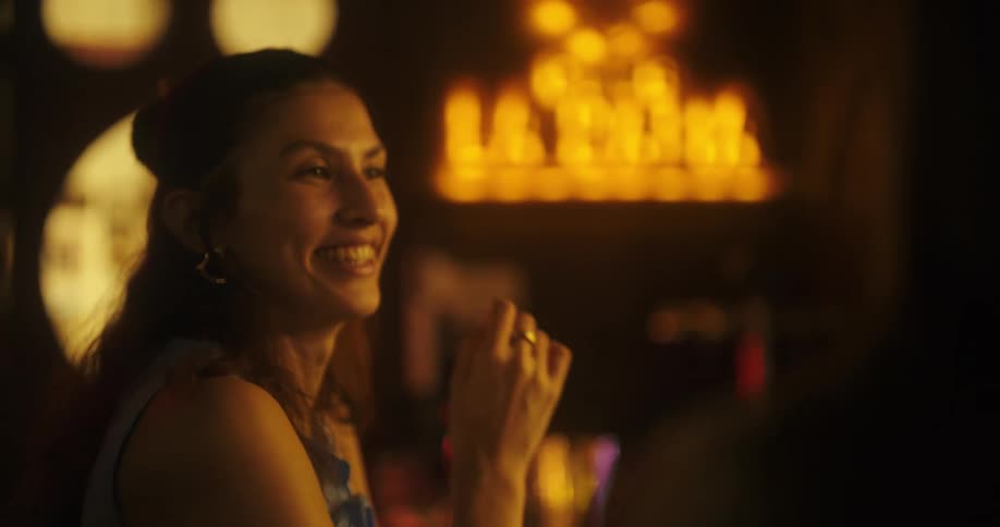 A woman with her hair in a bun smiles and laughs as she sips a drink with a blurred warm toned background