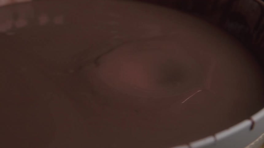 A dark chocolate colored liquid is contained in a large round vat and a piece of chocolate drops into the liquid from above causing a splash