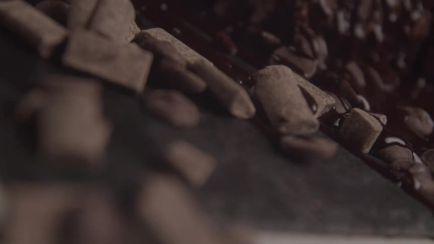 A chocolate factory conveyor belt is shown in a closeup shot as it carries a mixture of cubed and cylindrical chocolate pieces which are then coated in a rich dark liquid chocolate