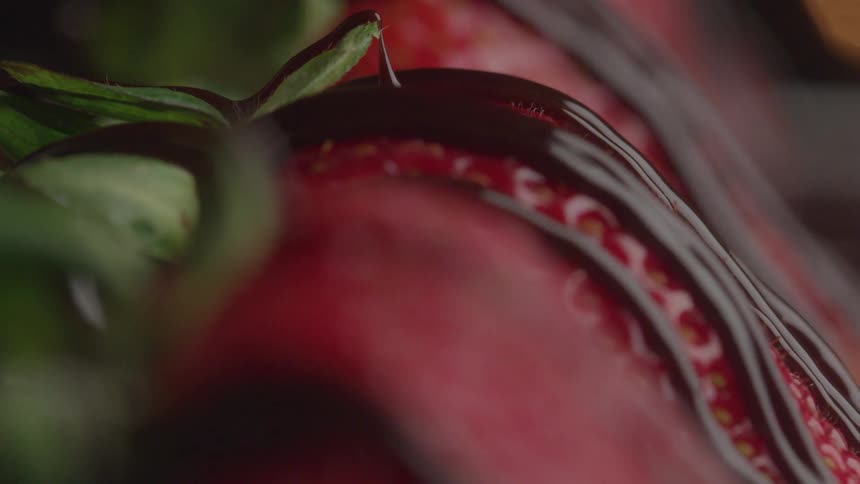 A close side view shows a red strawberry with visible white seeds being slowly drizzled with thick dark chocolate sauce