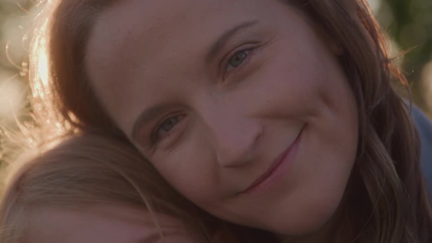 The video shows a closeup shot of two people embracing outside with the bright sun flaring in the background one person rests their head on the others shoulder and looks into the camera smiling warmly