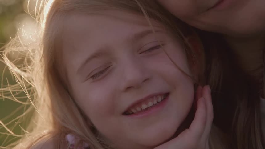 The video shows a closeup shot of a young girl with blonde hair who is smiling she is affectionately leaning against someone and the warm sunlight is flaring behind her creating a soft ethereal glow