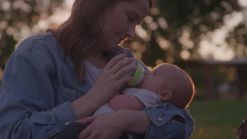 A mother is gently holding and bottlefeeding her baby outdoors illuminated by the soft warm light of the setting sun