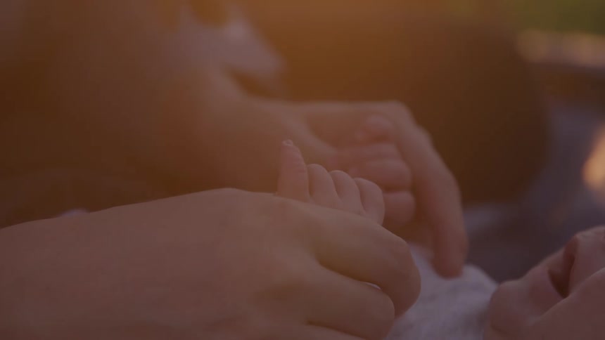 A womans hands holding a babys hands as the baby is lying on a blanket on the ground and the womans index fingers gently caress the babys chest