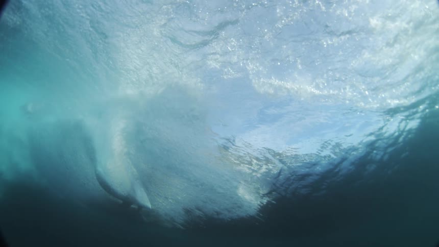 The video shows a submerged camera capturing a large wave from an underwater perspective the water is a clear turquoise blue and light ripples across the surface as the wave curls and crests overhead the shot has a somewhat distorted look due to the